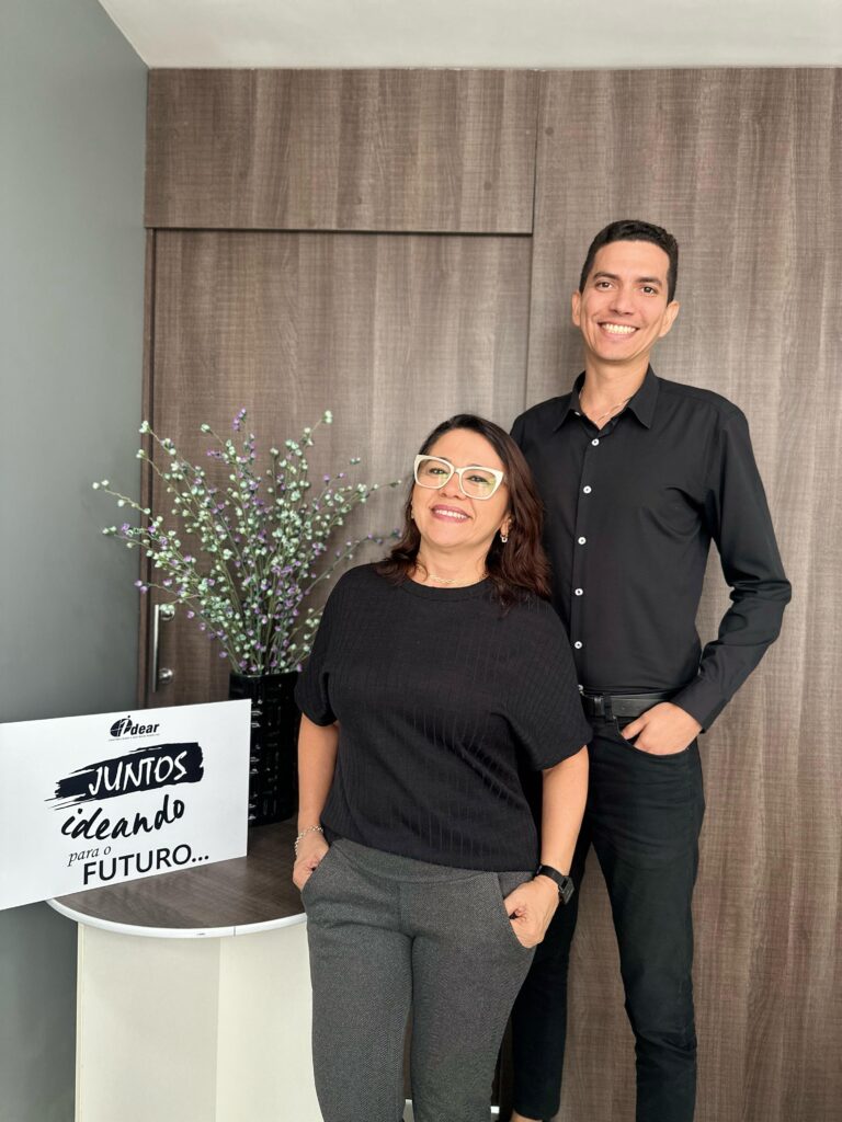 Foto dos diretores Waléria Leuda e Pedro Menezes, do Grupo Idear. Dois diretores estão em pé, um homem e uma mulher sorrindo com uma placa ao lado escrito "juntos ideando para o futuro".