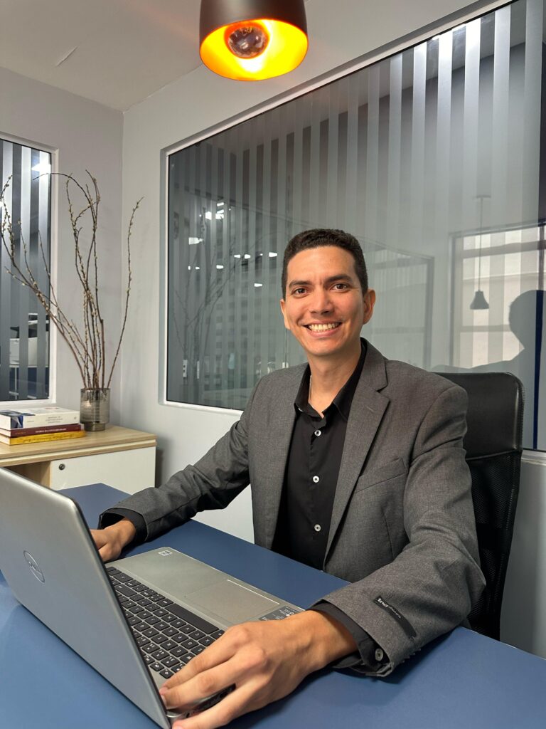 Diretor do Grupo Idear, Pedro Menezes. Na imagem um homem está sentado sorrindo utilizando um notebook.