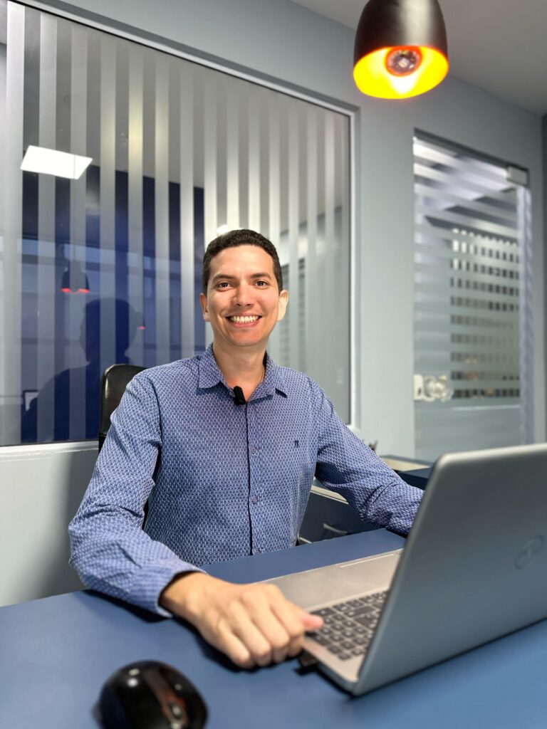 Diretor do Grupo Idear, Pedro Menezes. Na imagem um homem está sentado sorrindo utilizando um notebook.