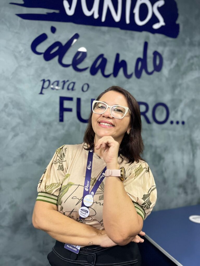 Waléria Leuda, diretora do Grupo Idear. Na imagem uma mulher está em pé sorrindo com a frase "Ideando para o futuro" na parede.